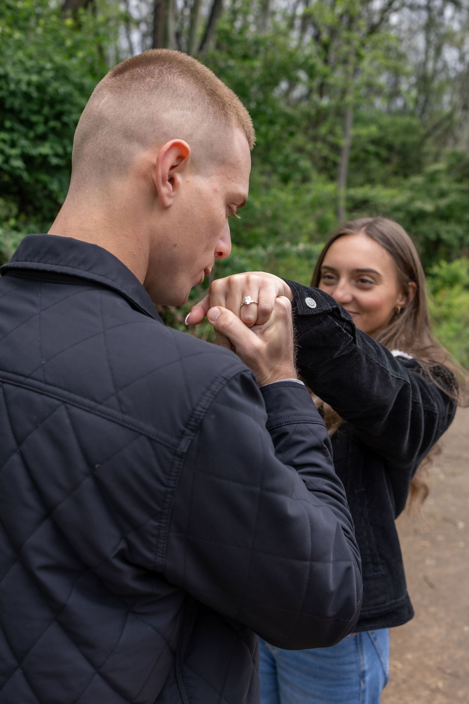 Man kissing woman's hand with engagement ring outdoors in a green park setting.