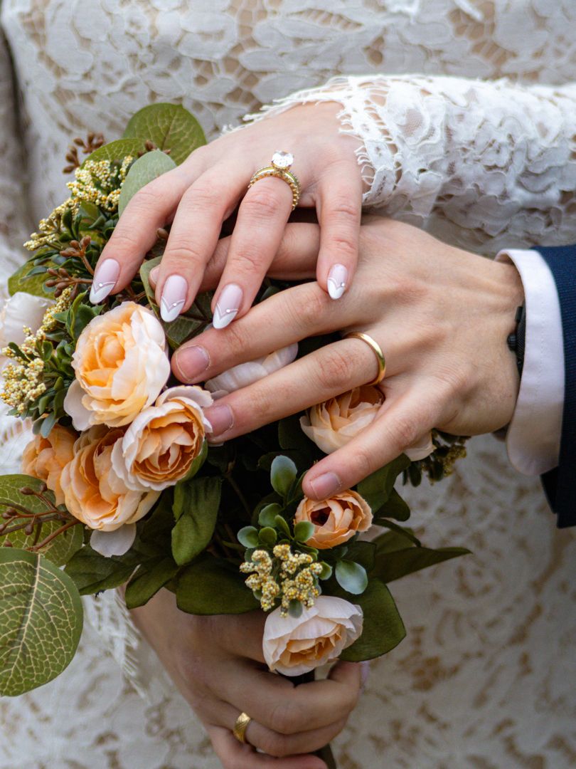 Bride and groom hands with gold wedding rings resting on a bouquet of peach roses and white flowers.