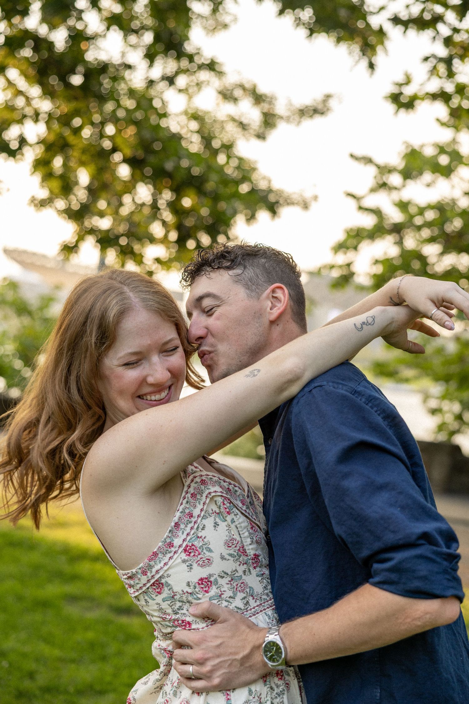 A joyful couple embraces outdoors, man kissing woman's arm, she laughs in floral dress, green trees behind them.