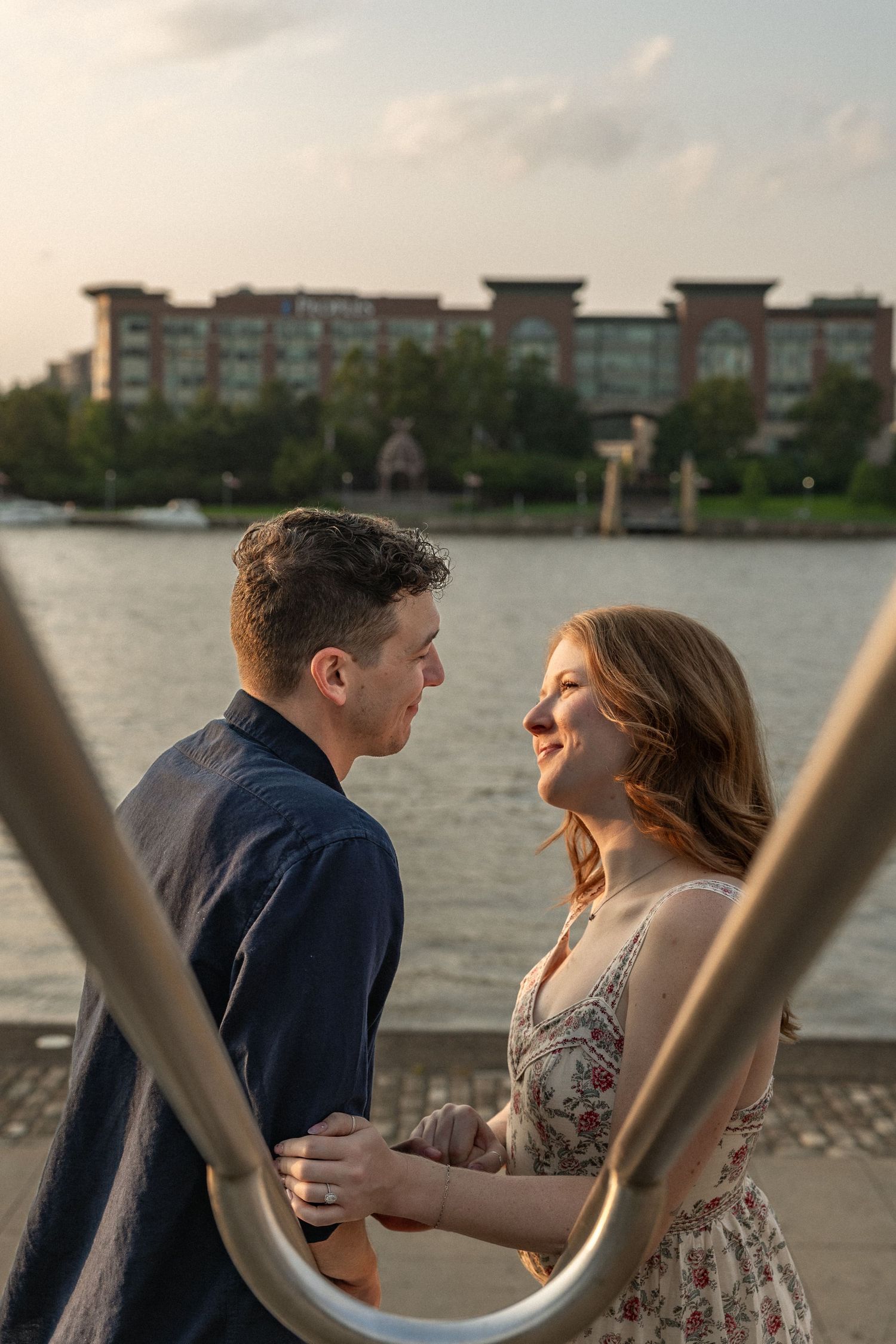 Couple holding hands and gazing at each other on a waterfront dock at sunset with a brick building across the river.