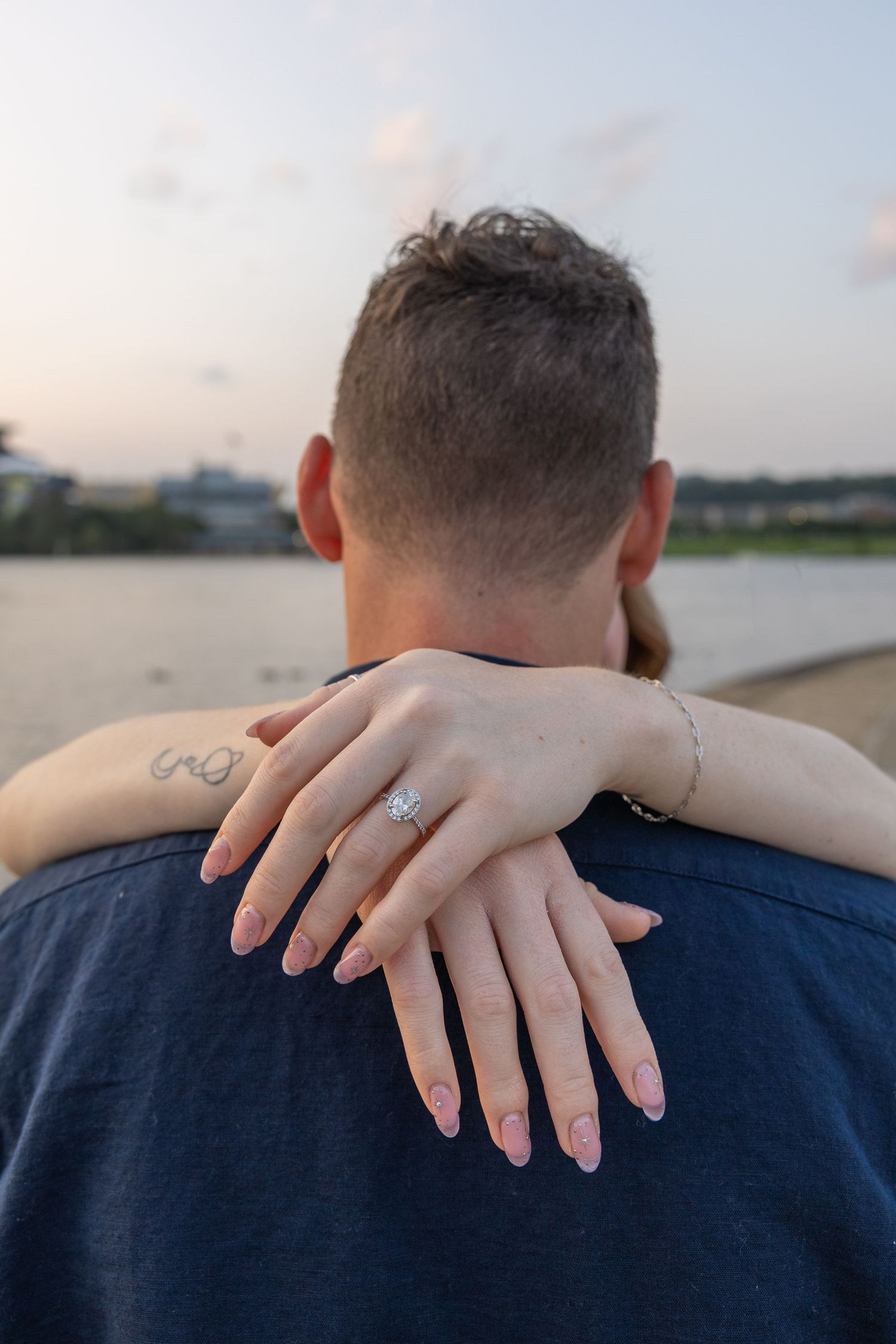 Couple embracing outdoors, woman's hand showing diamond engagement ring on man's shoulder near waterfront at sunset.