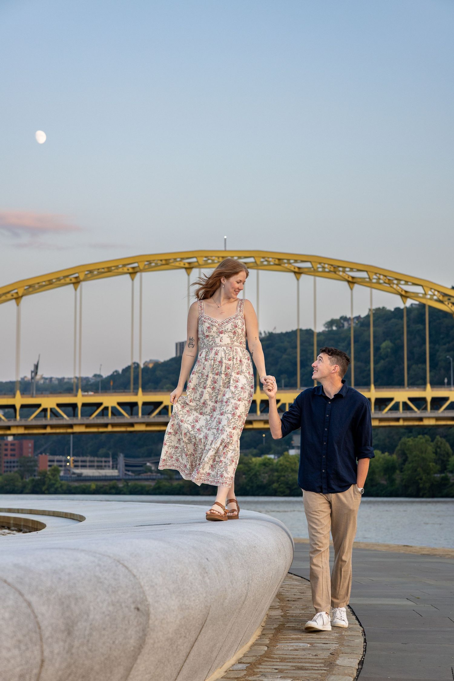 Couple holding hands near a river with a yellow arch bridge backdrop at dusk in Pittsburgh.