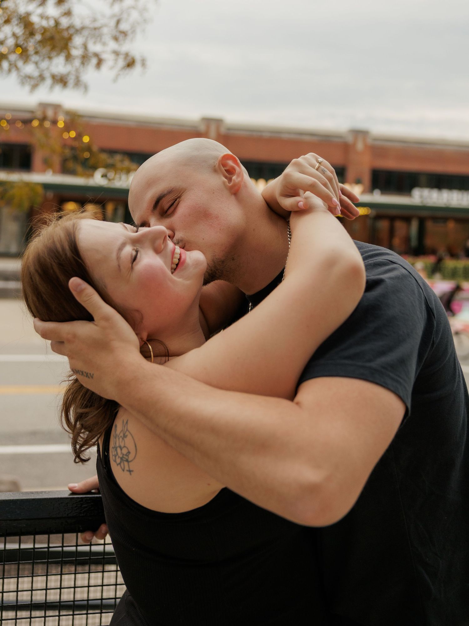 A bald man in a black t-shirt kisses a smiling woman with auburn hair as they embrace outdoors near a brick building.