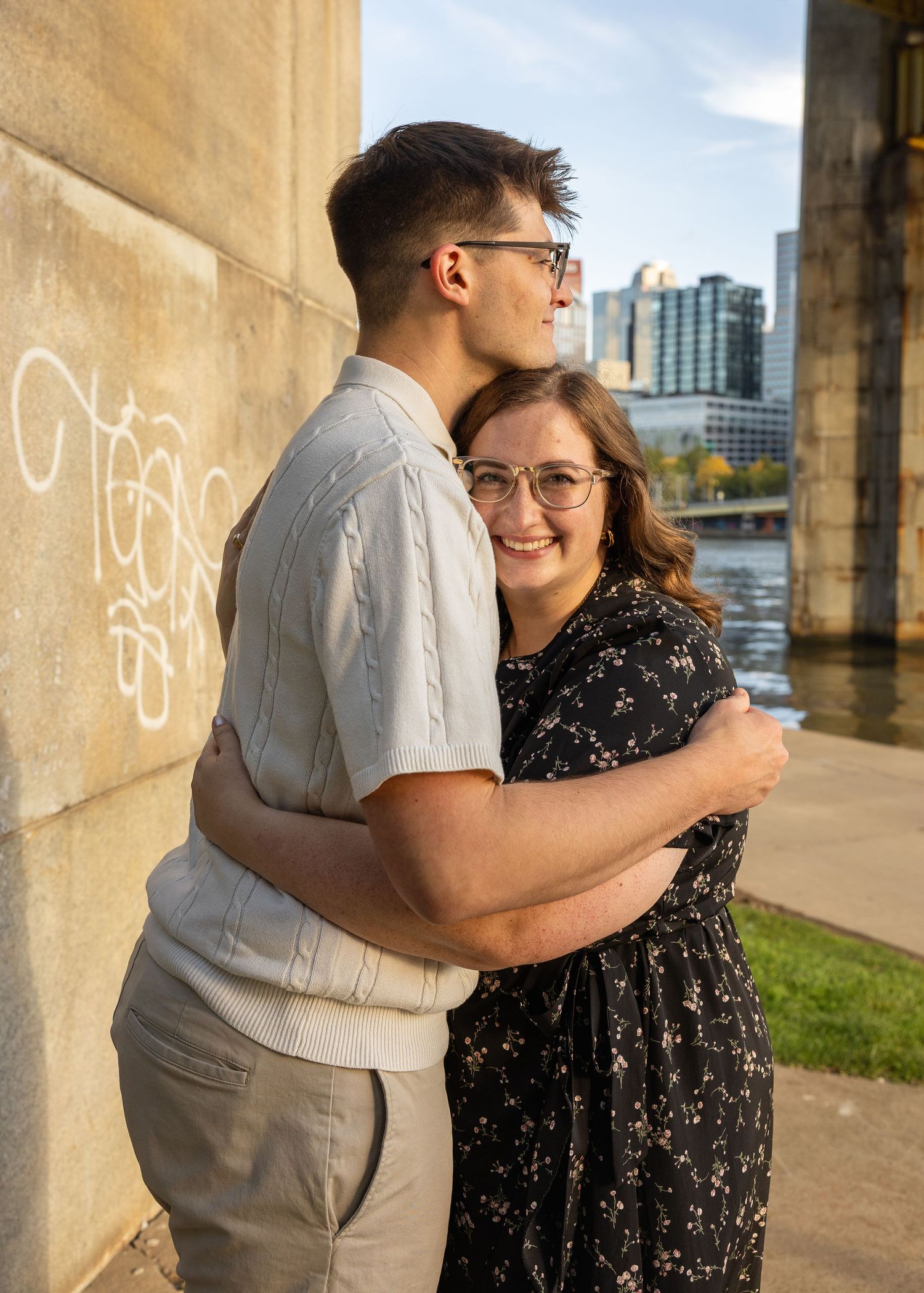 Smiling couple in glasses embracing under a concrete bridge beside a river, with city skyline buildings in the background.