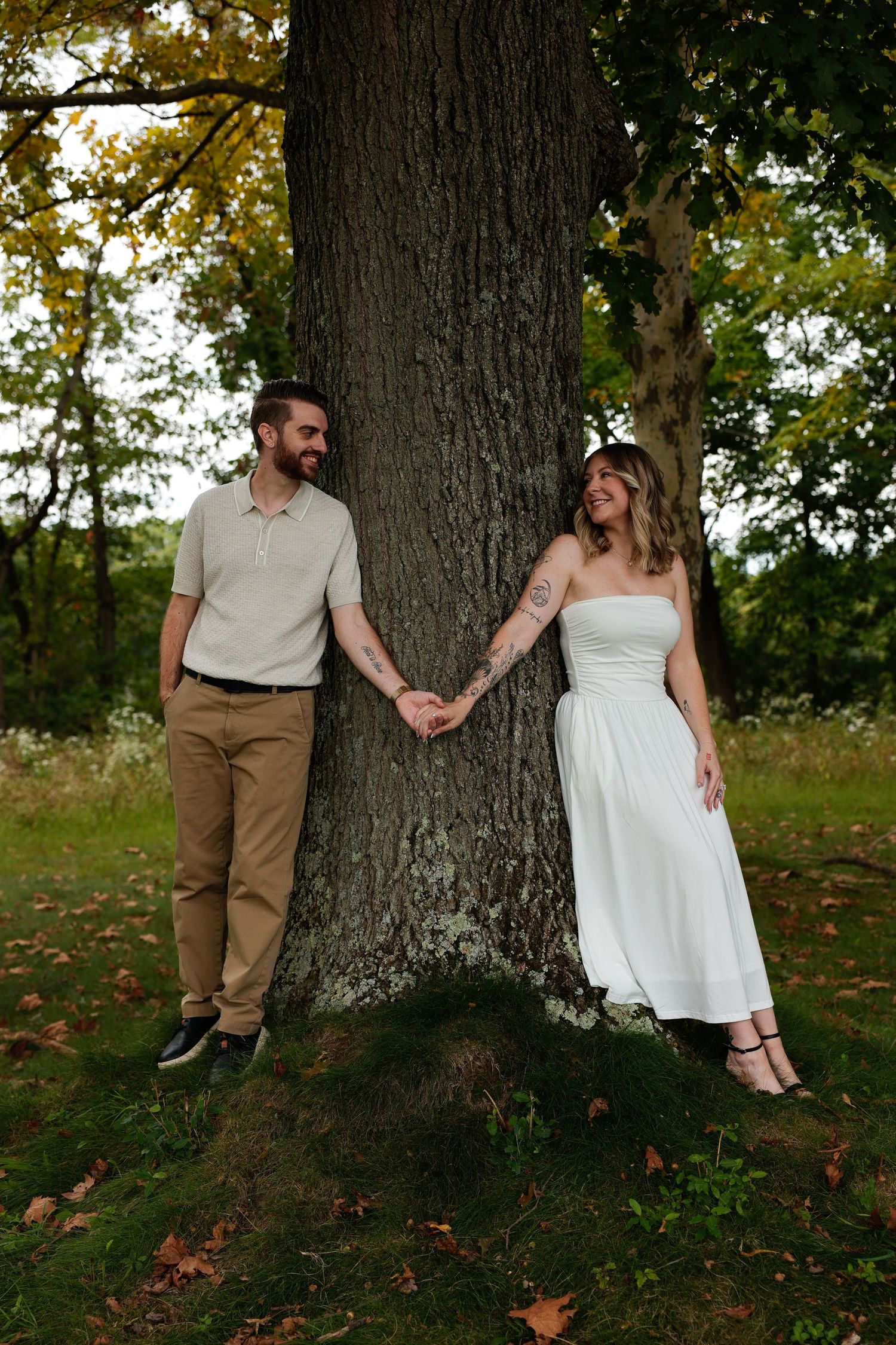 A couple holds hands behind a large tree trunk in a lush green park, smiling at each other.
