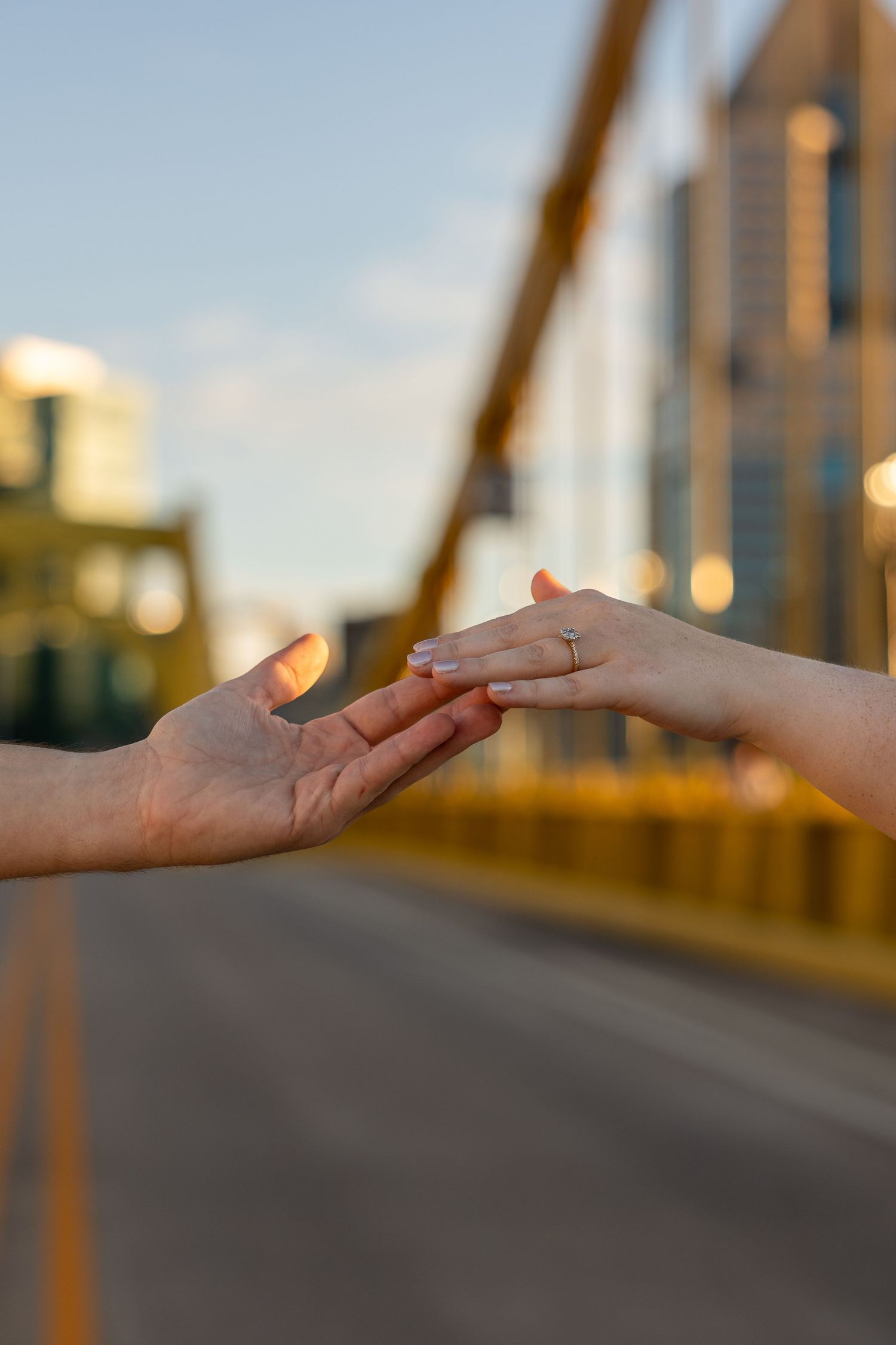 Two hands reaching toward each other on a golden-lit bridge at sunset, one wearing a ring, with a blurred city skyline behind.