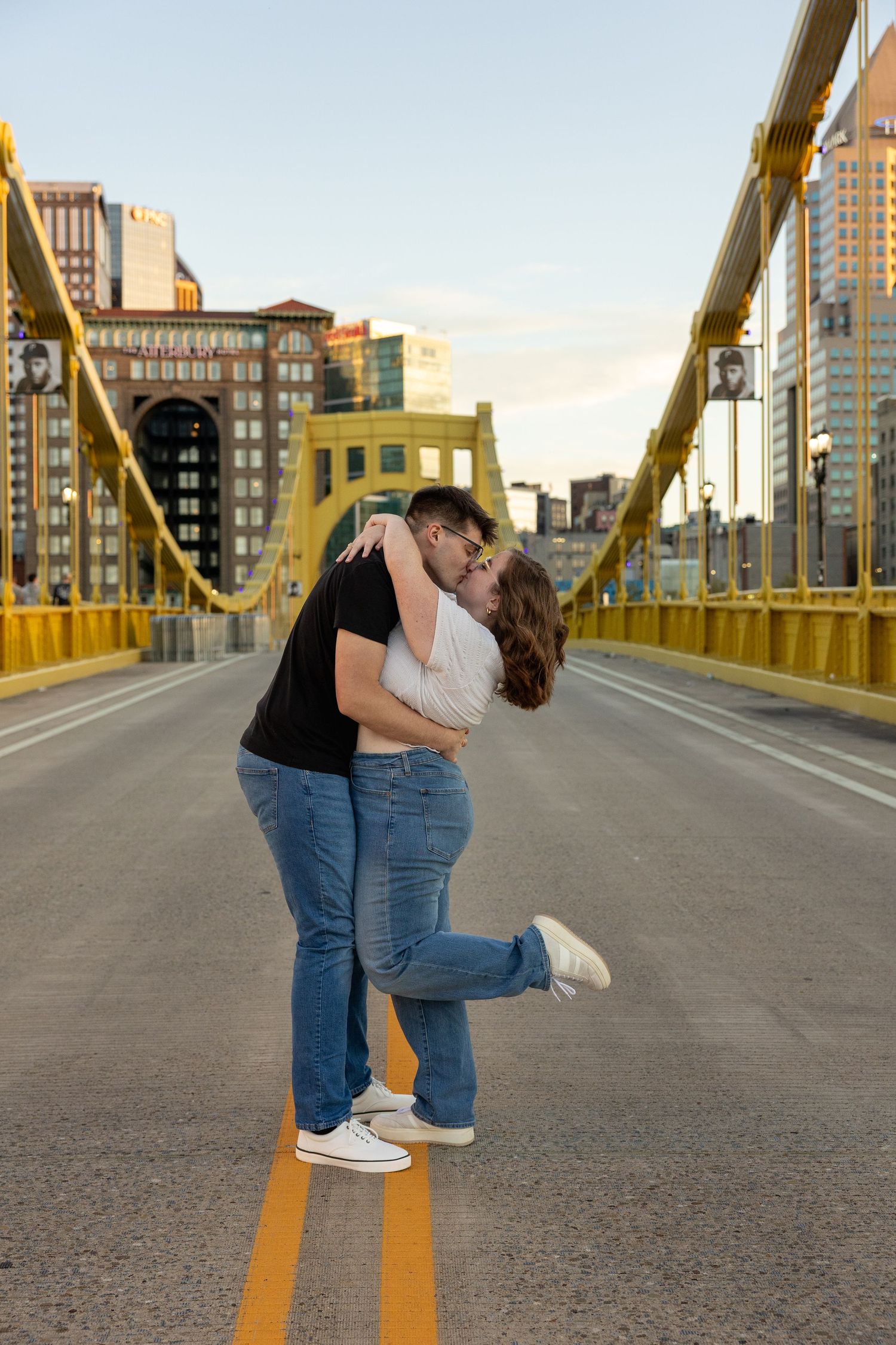 Couple in jeans and sneakers share a kiss on a yellow suspension bridge with city buildings behind them at sunset.
