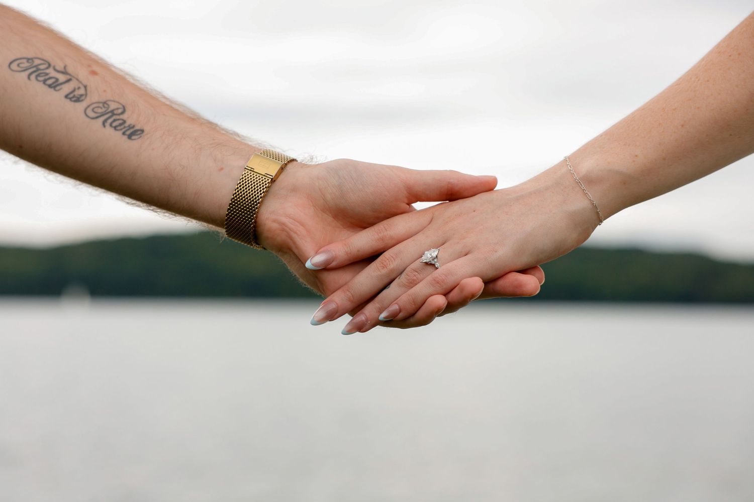 Close-up of couple holding hands showing engagement ring and tattoo with lake and trees in background.