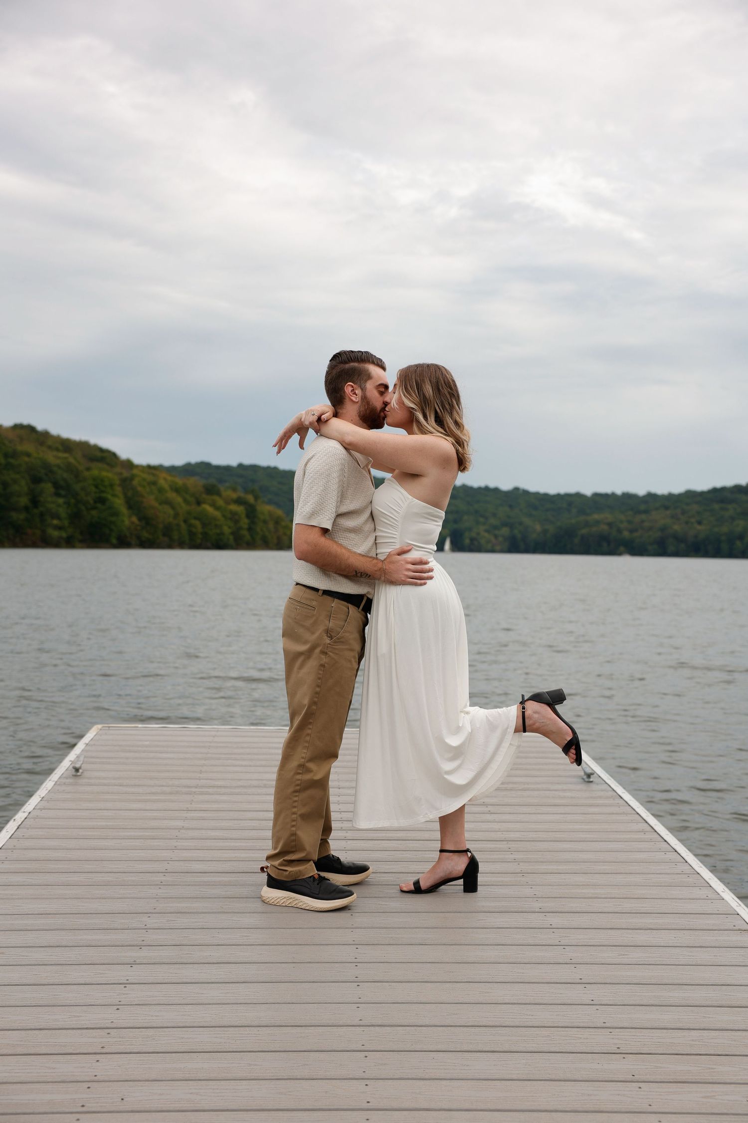 Couple sharing a romantic kiss on a lakeside dock, woman in white dress lifting her heel, surrounded by serene water and trees.
