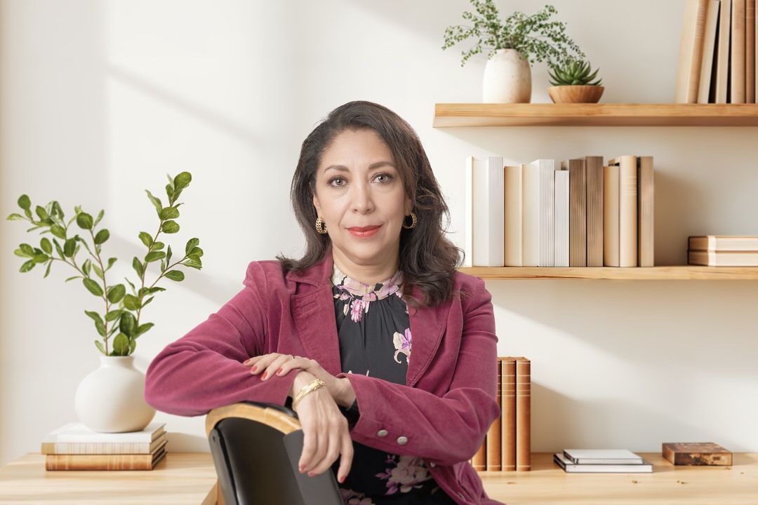 Retrato profesional de mujer con blazer rosa en ambiente de home office iluminado y estantería decorativa.