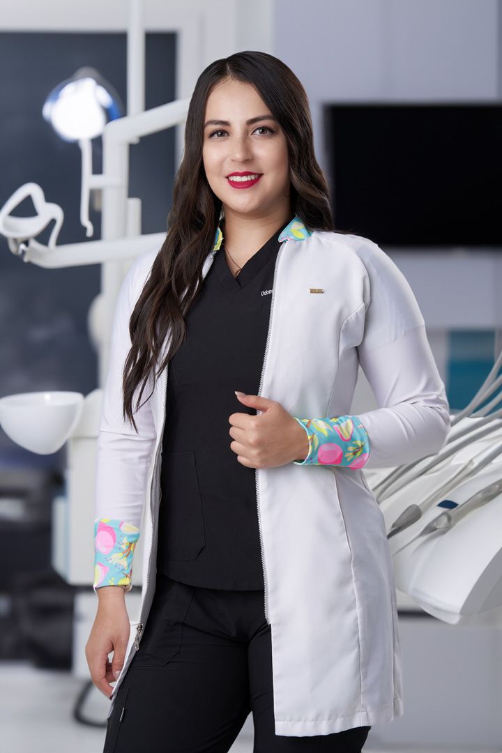 Retrato profesional de dentista u odontóloga con bata médica en su consultorio con equipo dental al fondo.