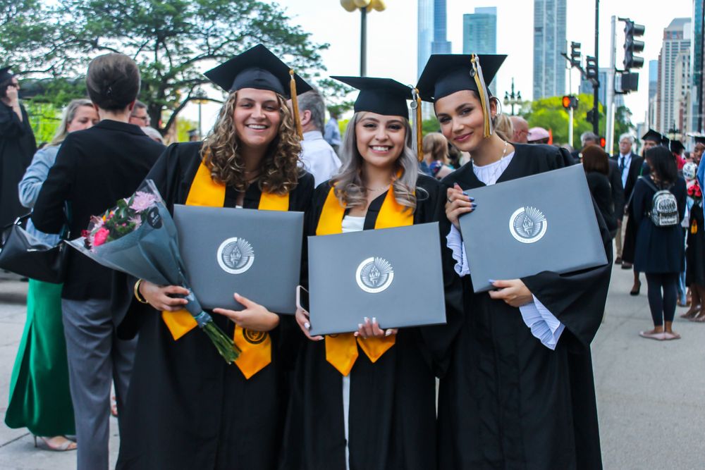 Group of graduates in caps and gowns holding diplomas with city skyline in background.
