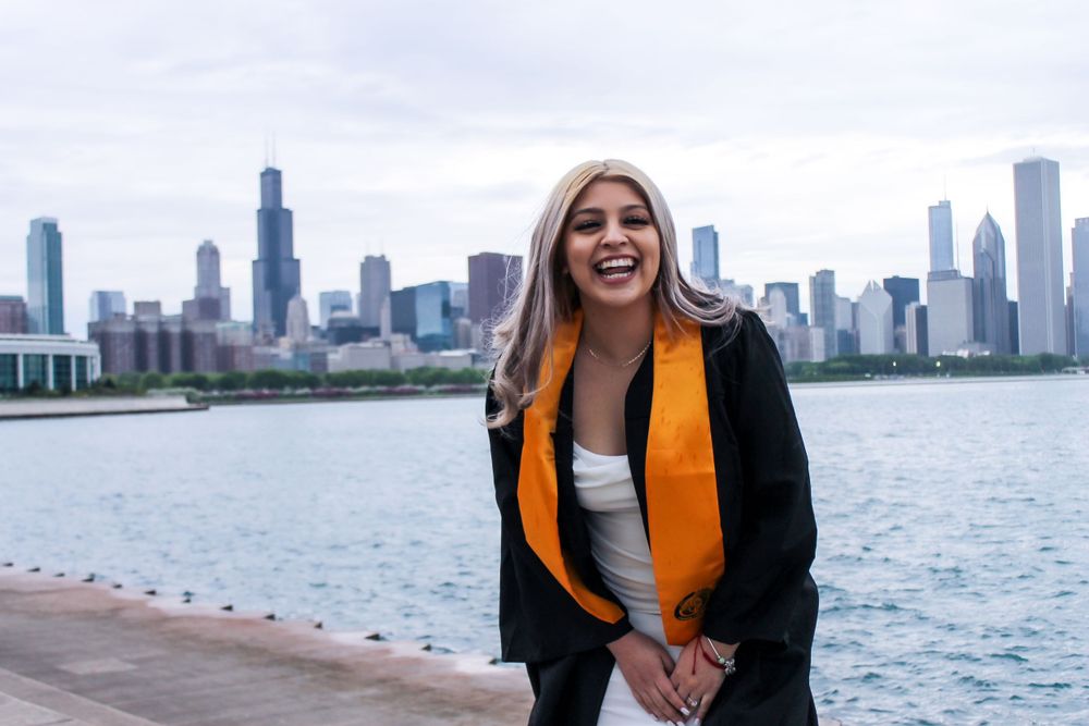 Graduate posing in a white dress and orange stole against a city skyline backdrop near the water.