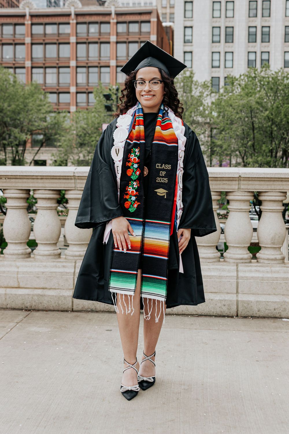 Graduate wearing traditional graduation regalia poses on stone balcony overlooking city.