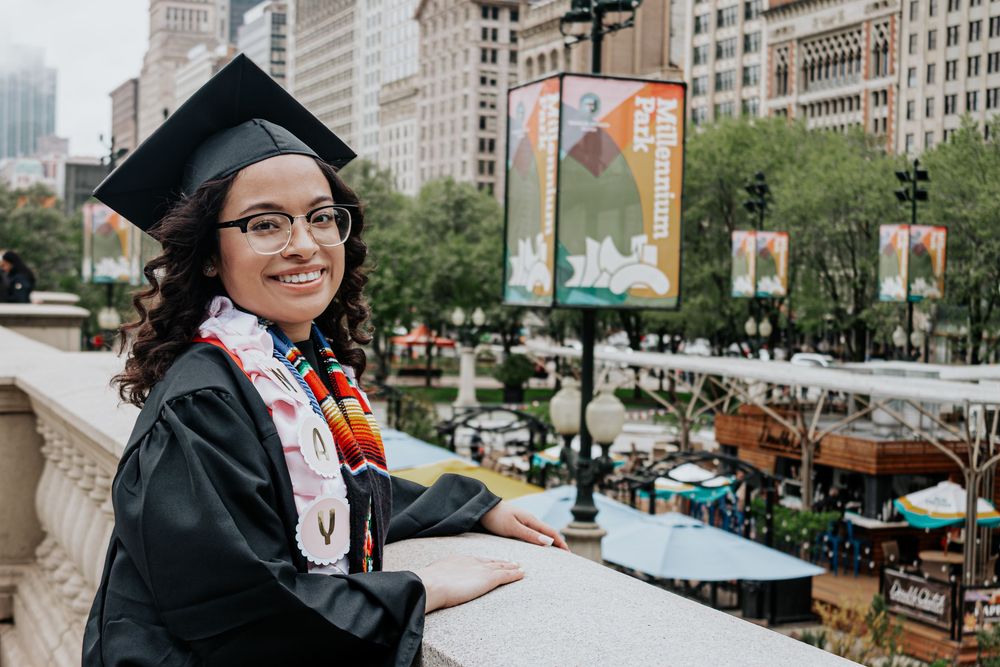 Graduate poses with diploma in front of urban cityscape.