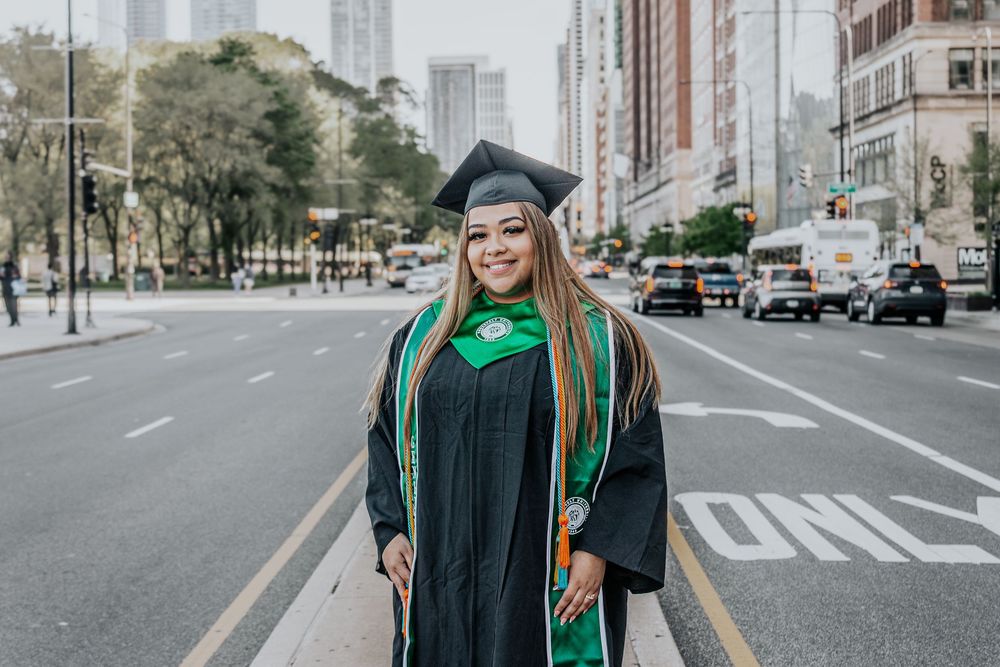 Graduate in cap and gown poses on city street with urban buildings in background.