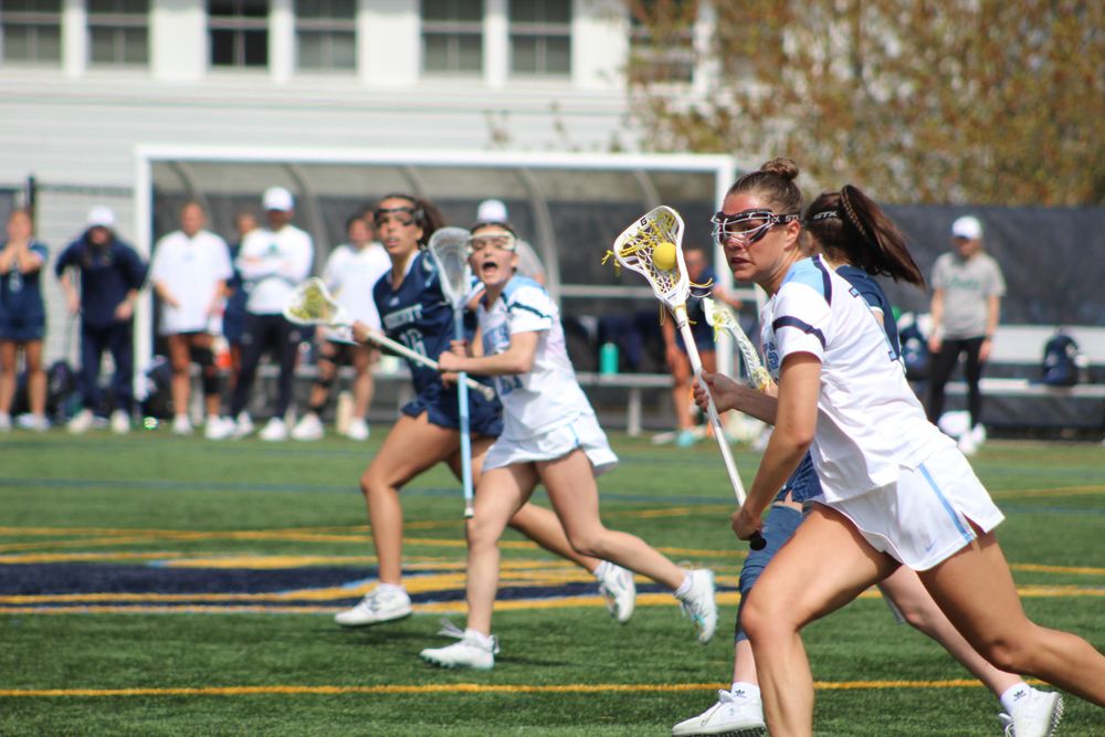 Lacrosse players in white and navy uniforms sprint across field during competitive game.