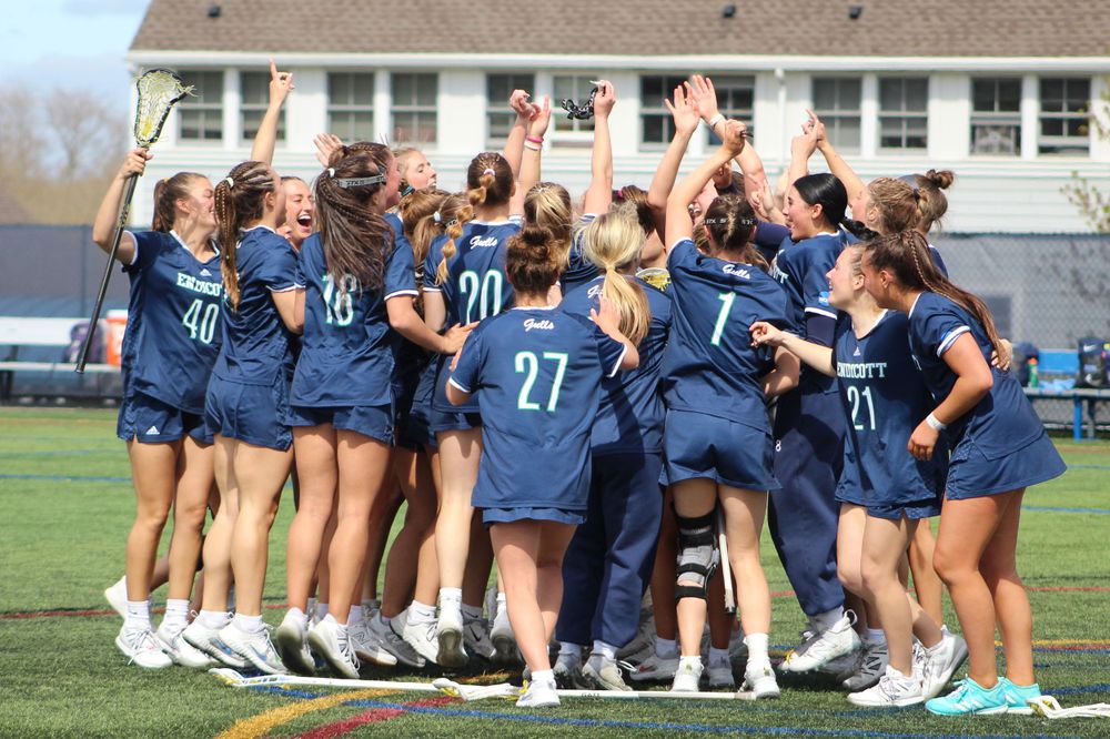 Women's lacrosse team celebrates in huddle wearing navy blue uniforms on sports field.