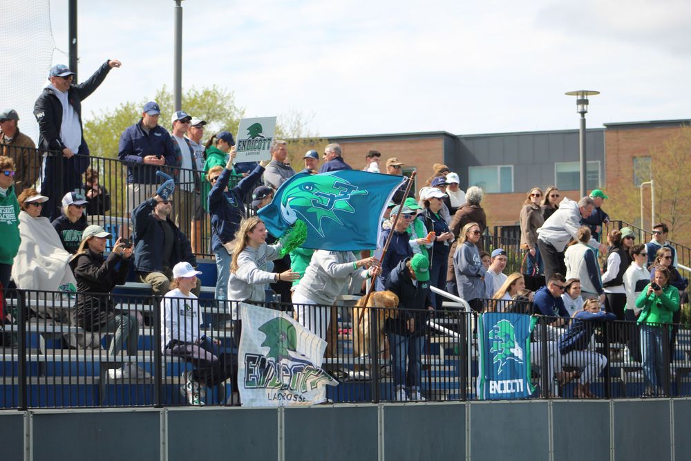 Energetic fans wave flags and cheer from bleacher section during outdoor sporting event.