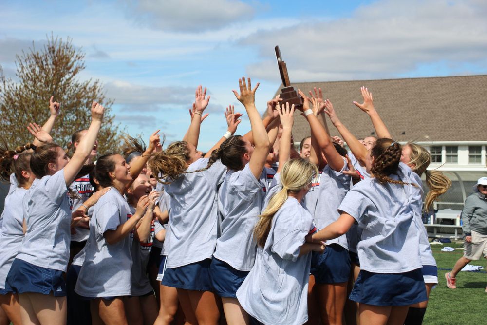 A women's sports team celebrates victory by raising their hands and sticks in the air on an outdoor field.