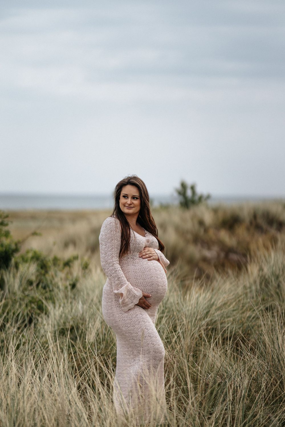 A person in a lace maternity dress poses gracefully amid tall grasses in a rugged outdoor landscape at sunset.