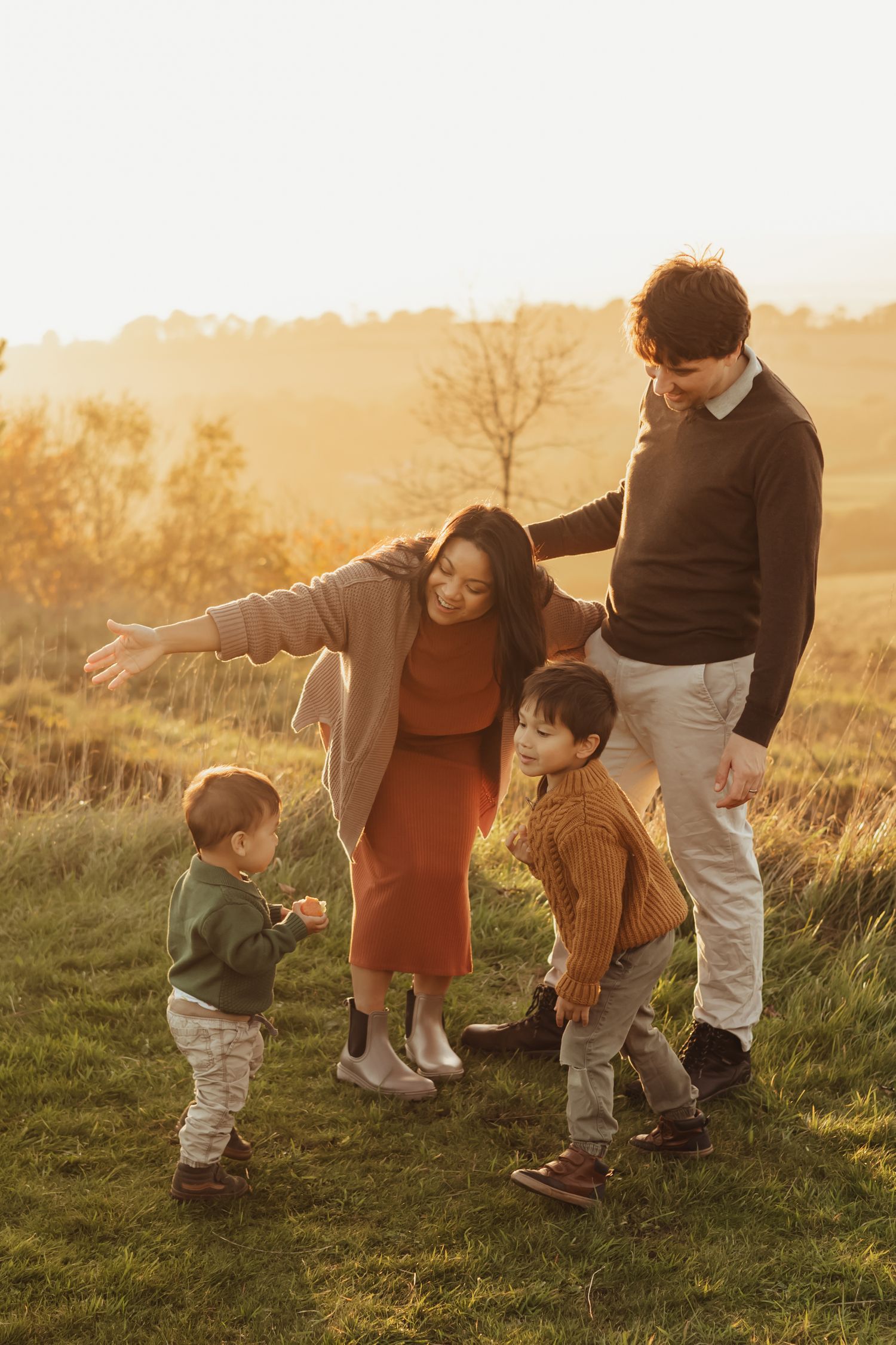 A family plays together outdoors during golden hour autumn light, swinging a child while laughing in amongst trees.