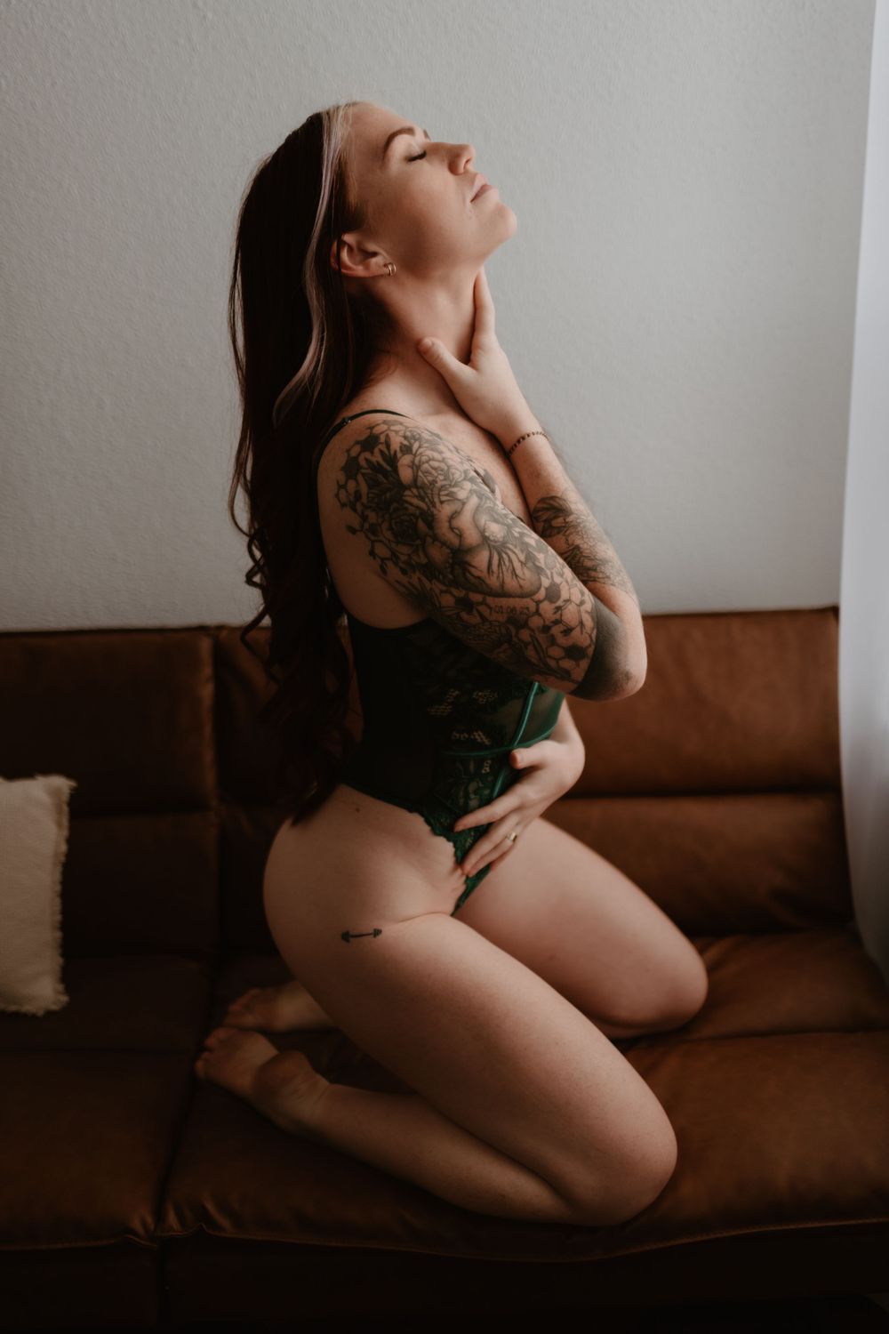 A person in a green bodysuit poses sensually on a brown couch, displaying sleeve tattoos in a moody boudoir setting.