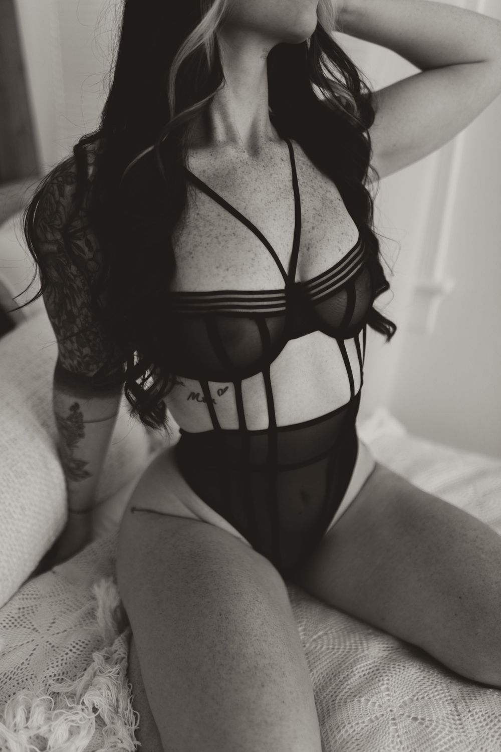 Black and white photo of someone in strappy lingerie posing on lace bedding.