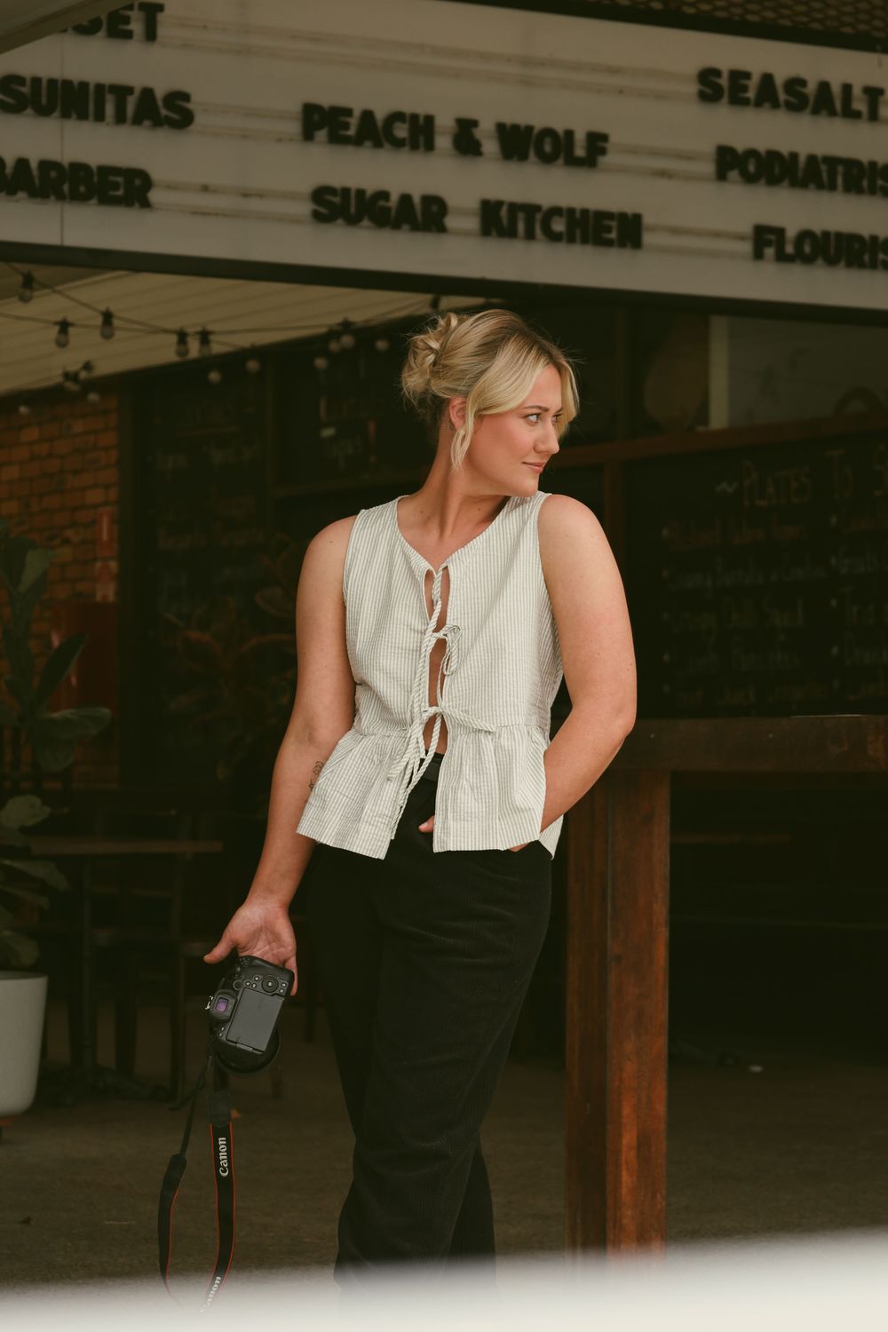 A figure in a white sleeveless top and black pants stands in a dimly lit interior space with business signage visible above.