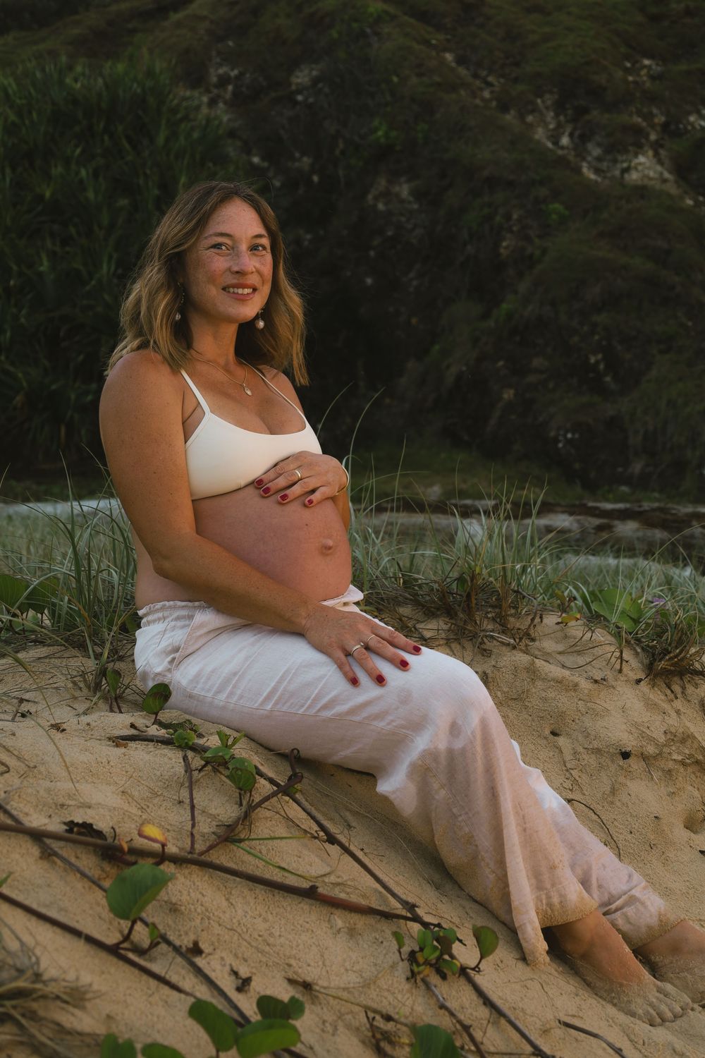 A person in a white dress sits on a sandy beach slope surrounded by greenery and natural scenery.