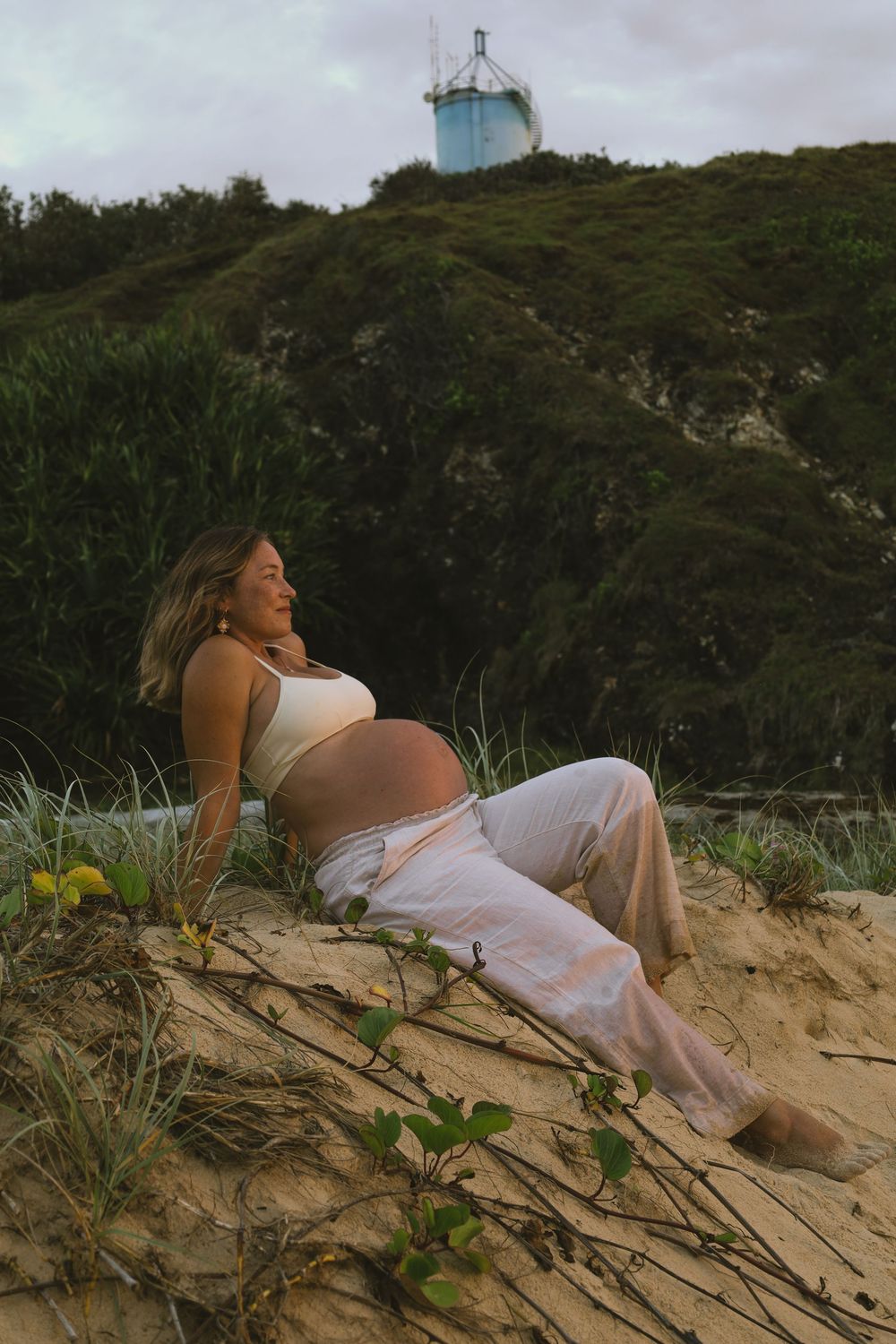 A pregnant woman in white dress reclines peacefully on a sandy beach dune surrounded by coastal vegetation.