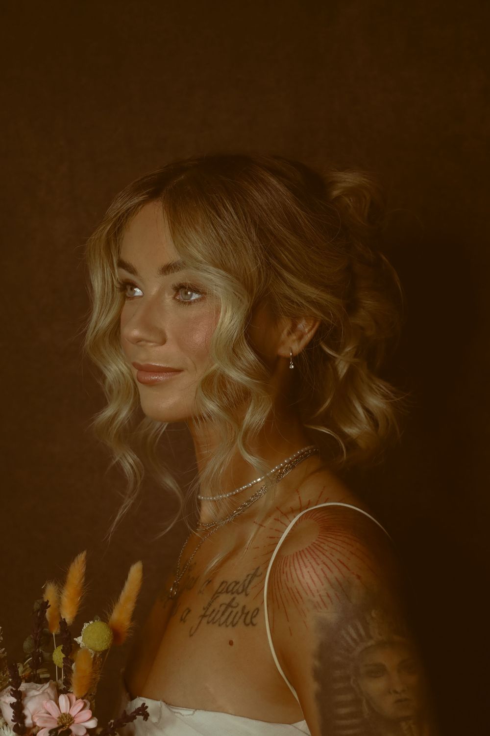 Artistic portrait in warm sepia tones features a woman with wavy hair wearing a delicate necklace and holding flowers.