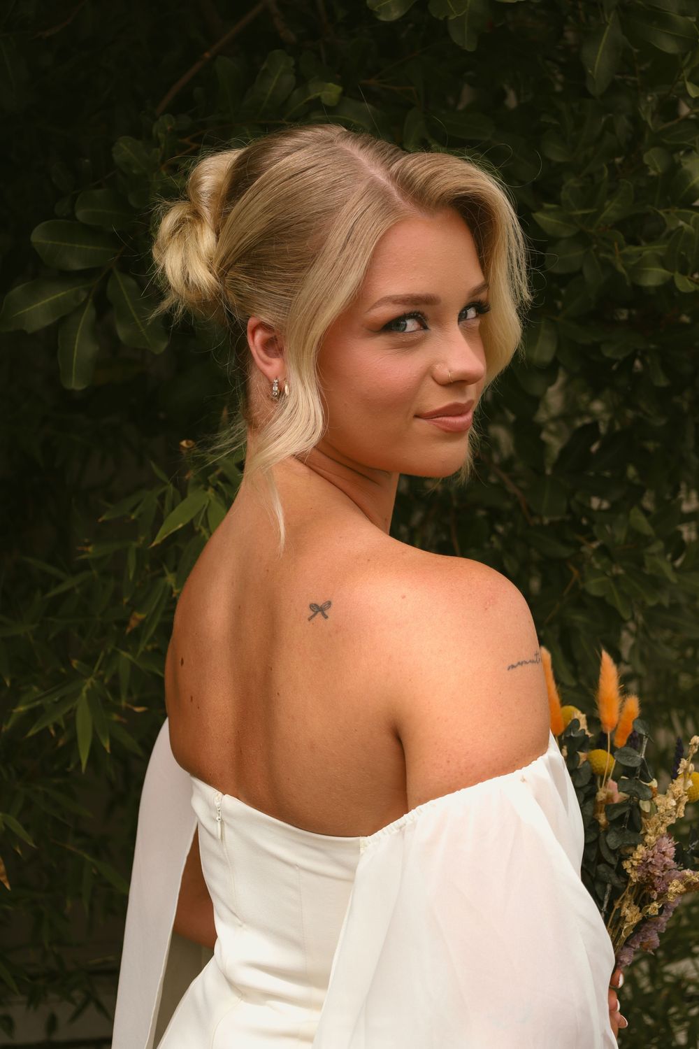 A woman in a white off-shoulder dress poses outdoors with a small butterfly tattoo visible on her shoulder.
