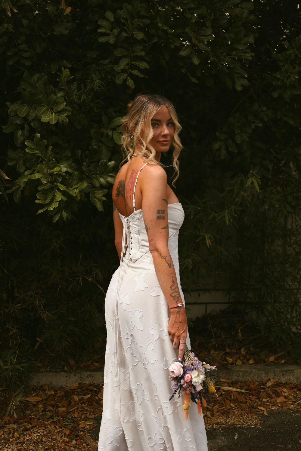 A person in a white lace wedding dress stands in a garden setting holding a bouquet against a dark foliage background.