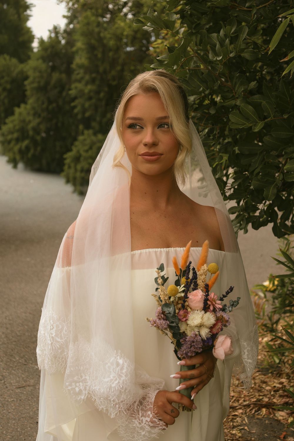A bride in a white dress with delicate lace veil holds a colorful wildflower bouquet against a leafy green backdrop.