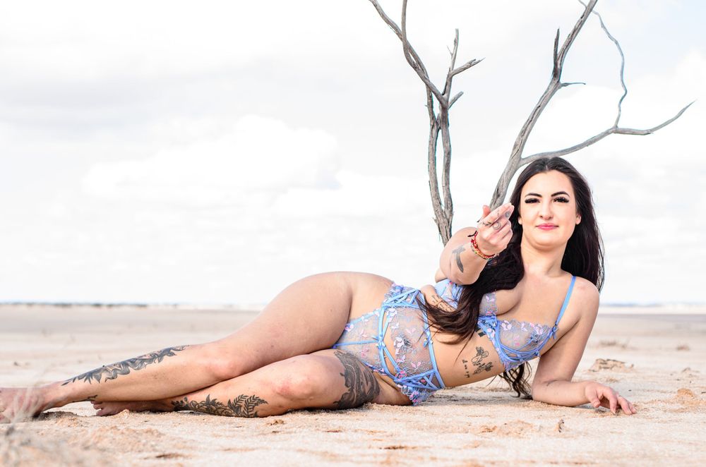 Person in blue lingerie set reclines on sandy ground near dried tree.