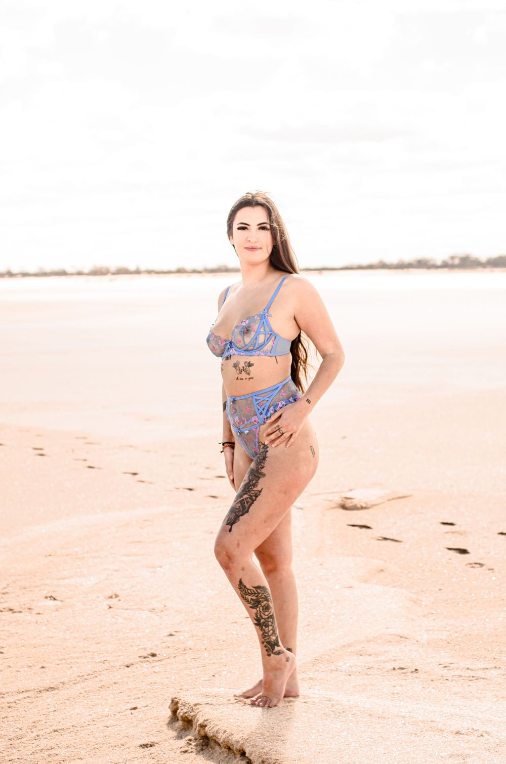Model with tattoos poses in blue swimwear on sandy beach.