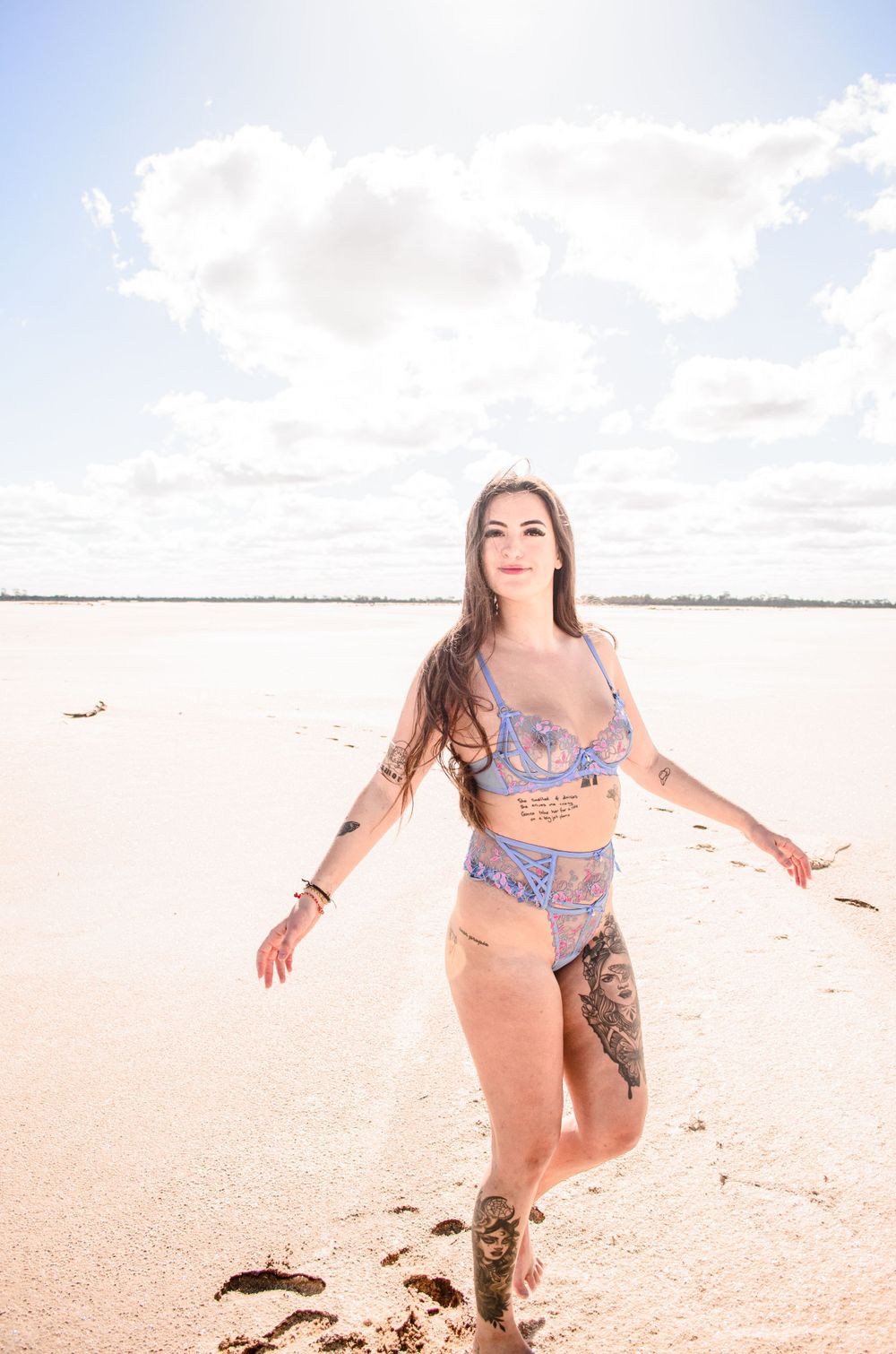 Model in blue two-piece swimsuit walks along beach under cloudy sky.