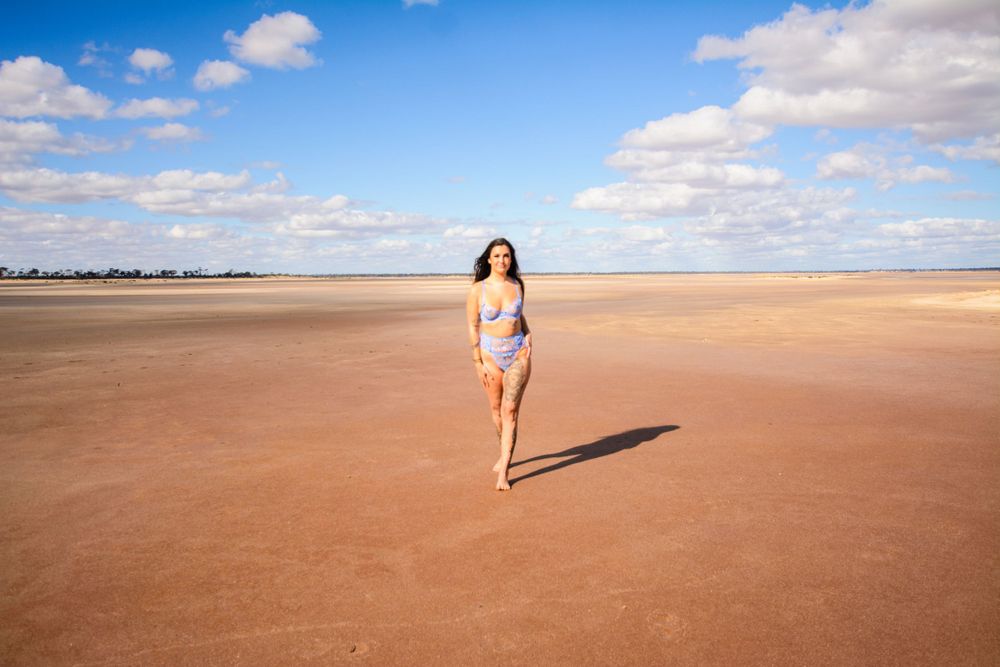 Model runs across pink sand beach wearing blue swimwear in bright sunlight.