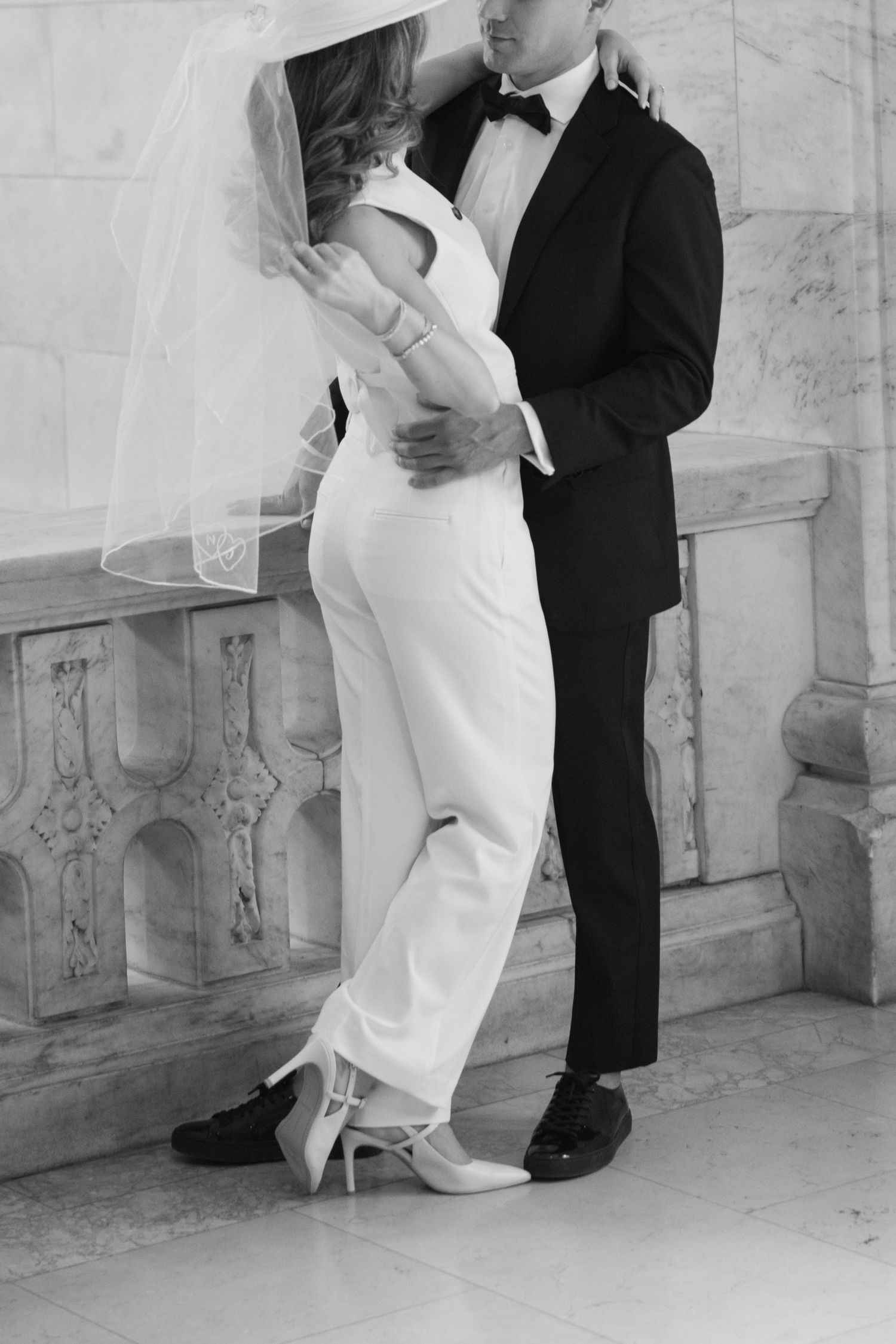 Black and white photo of a bride in white jumpsuit and groom in tuxedo embracing near marble columns.