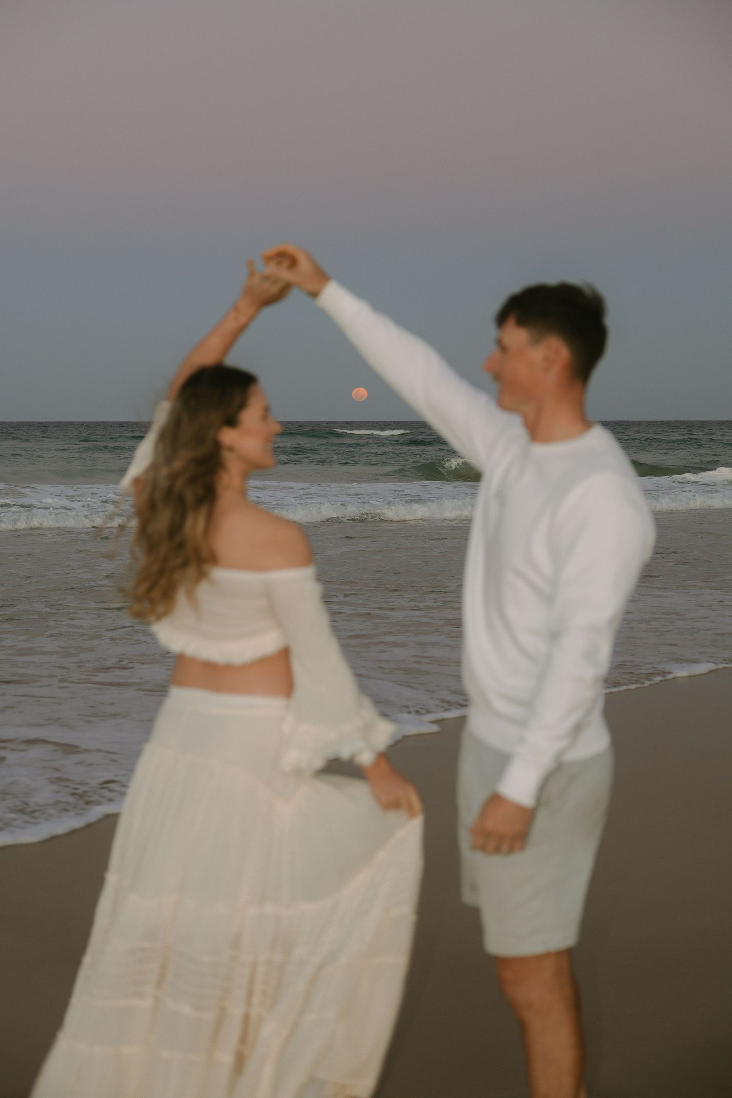 Couple dancing on a beach at dusk as the man twirls the woman in a flowing cream skirt beside the ocean waves.