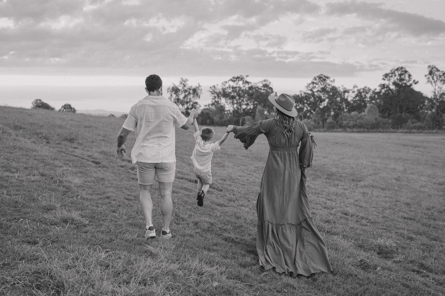 Black and white photo of parents walking hand in hand with their toddler across a grassy hillside under a cloudy sky.