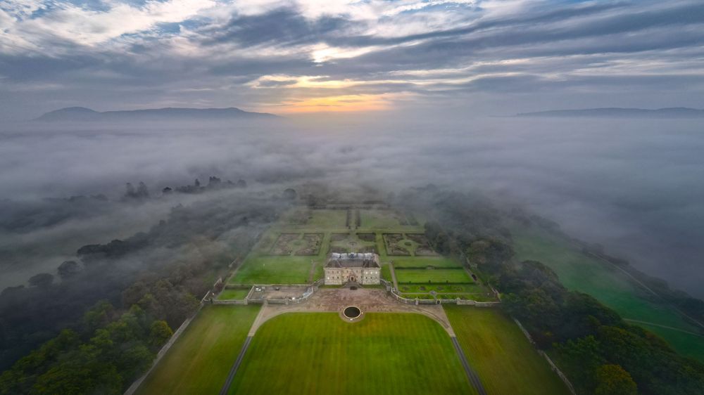 Misty morning aerial view over agricultural fields and mountains under dramatic cloudy skies.