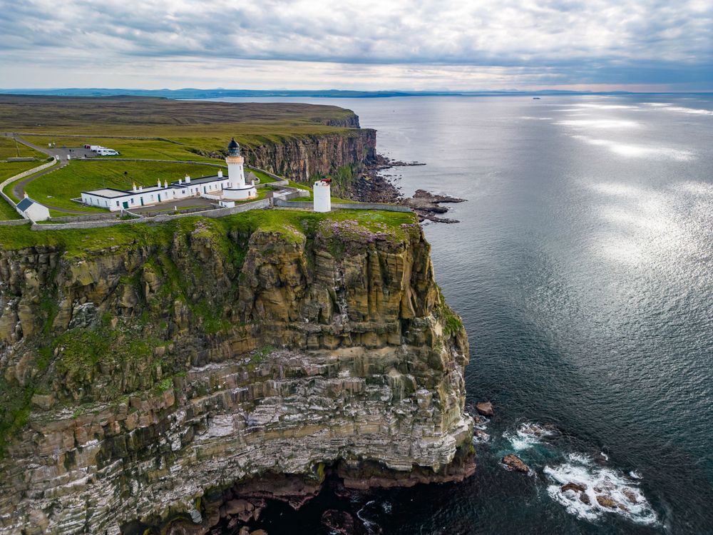 Panoramic aerial view of white lighthouse buildings situated on dramatic coastal cliffs above the ocean.