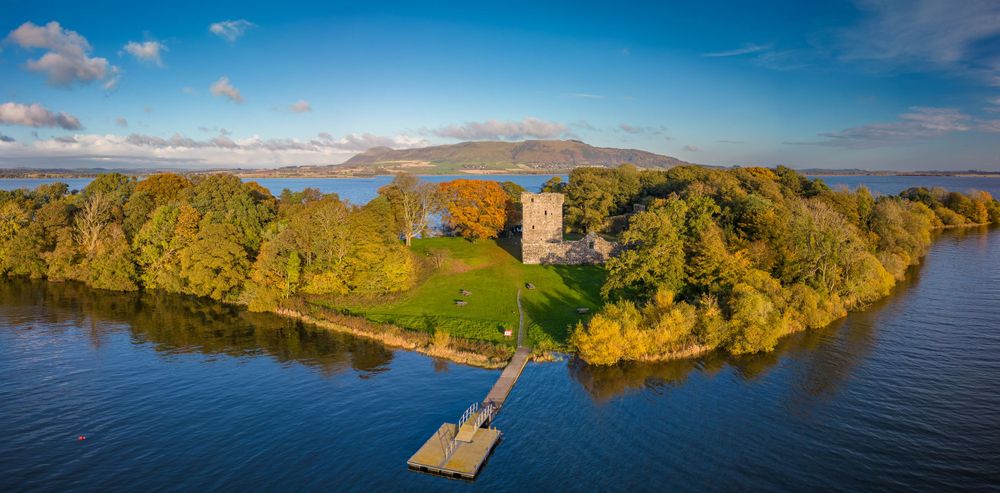 Wide aerial view of coastline with historic tower ruins and autumn trees beside blue waters.