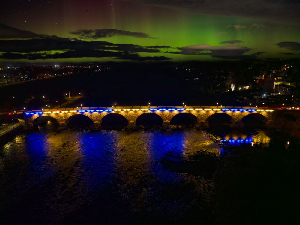 Northern lights illuminate the night sky over a bridge with colorful reflections in the water.