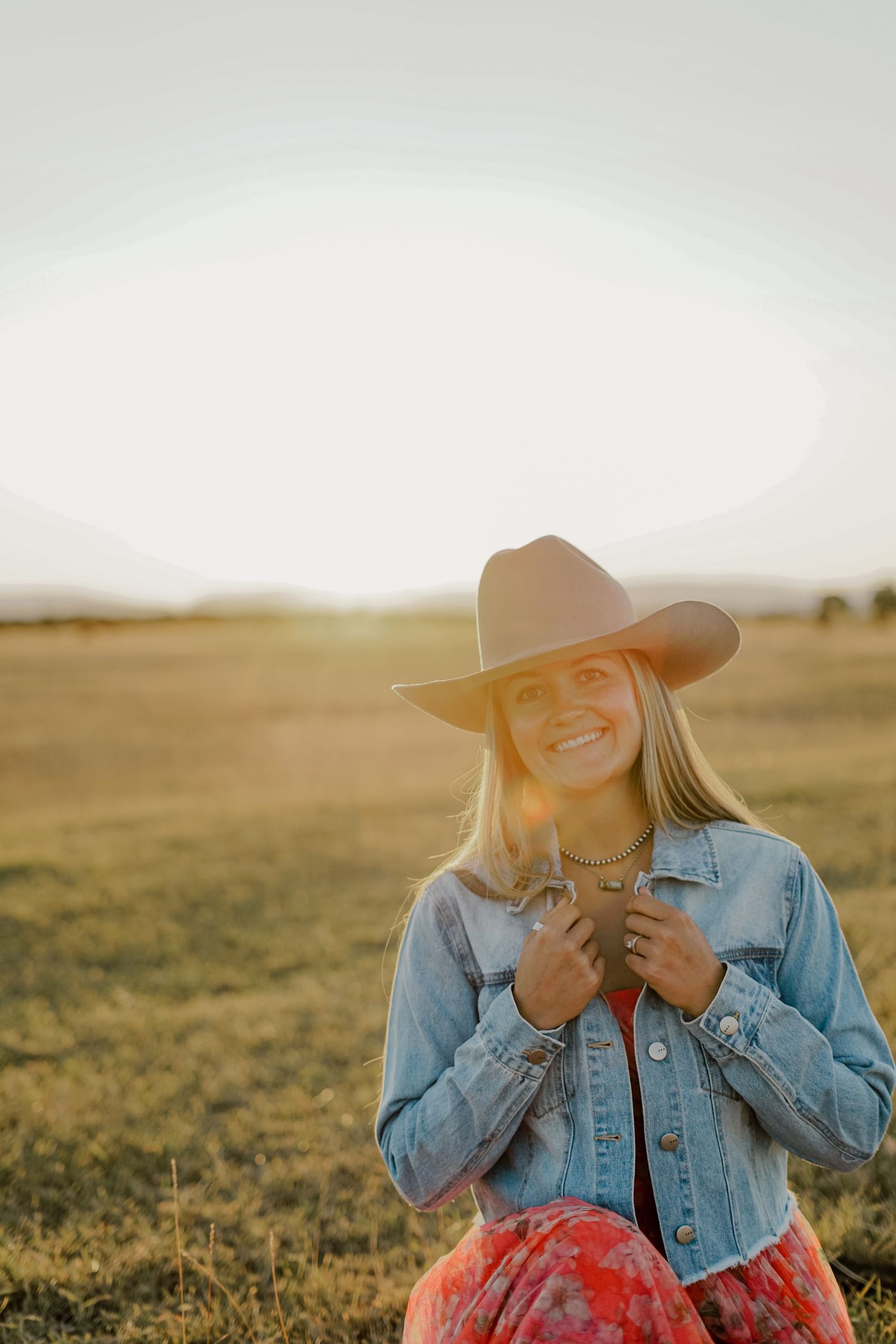 A woman in a denim jacket and cowboy hat smiling into the camera with sunset and mountain in the background