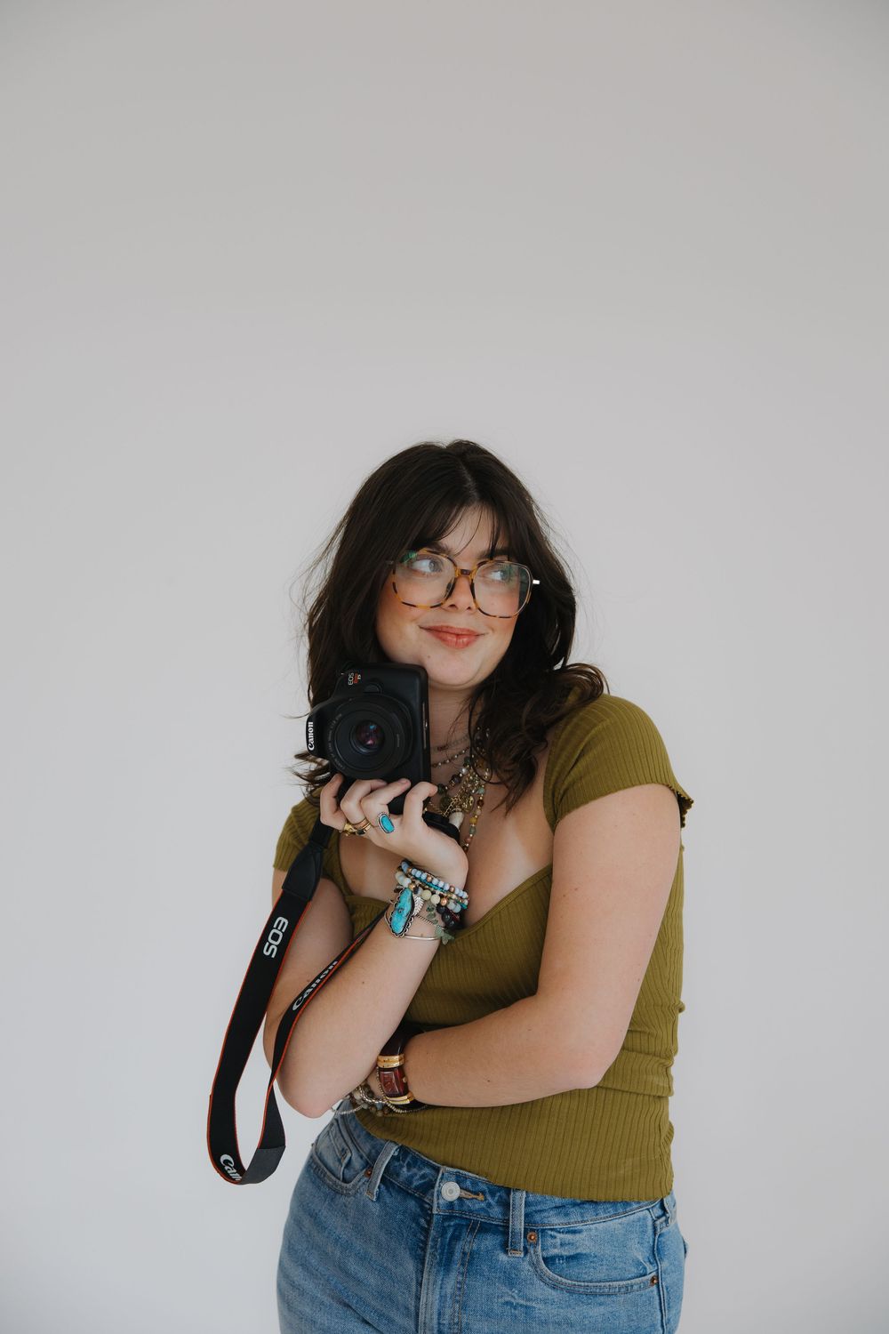 Photographer in olive green top and denim holds professional camera while wearing stylish glasses and jewelry against white wall.