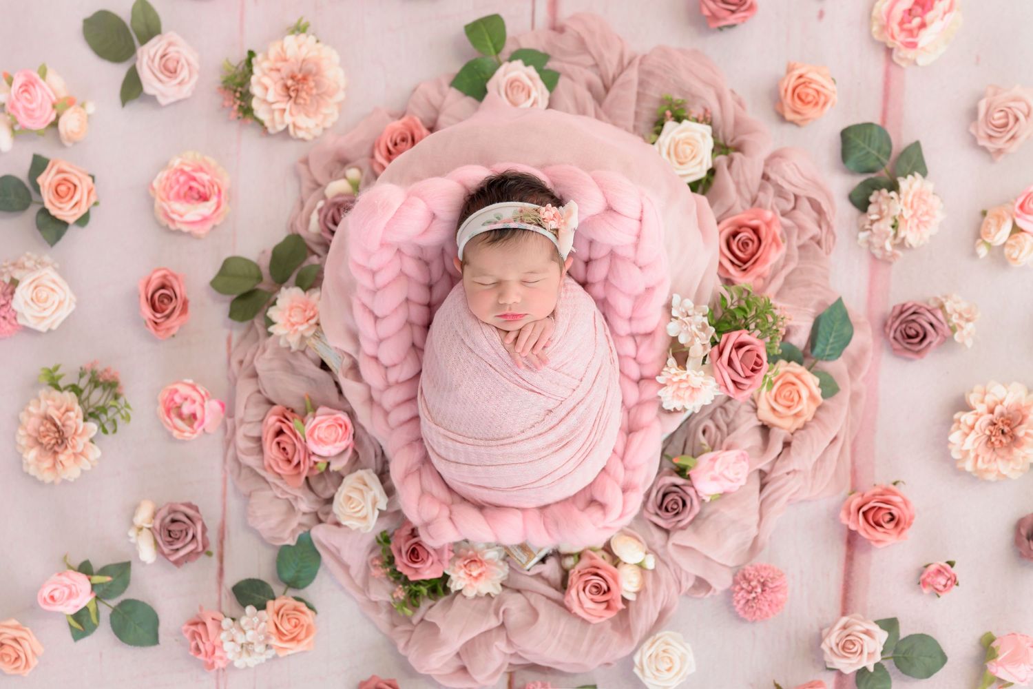 A peaceful newborn photography setup with soft pink swaddle and roses arranged around a heart-shaped basket.