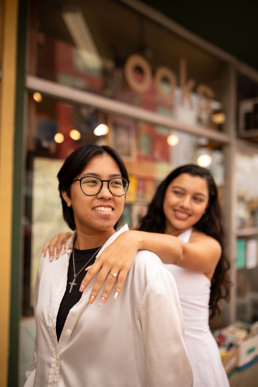 Two friends pose together outside a storefront at night, with one showing off an engagement ring.